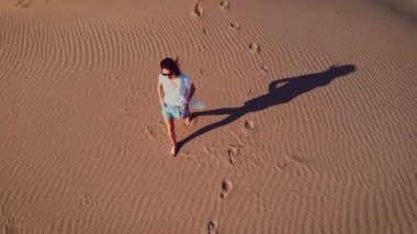Kadın, altın gün batımı ışığı ve rüzgarda, çölde dalgalı kum tepelerinde yürüyor..
