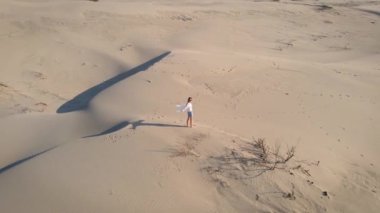 Kadın, altın gün batımı ışığı ve rüzgarda, çölde dalgalı kum tepelerinde yürüyor..