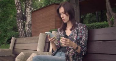 Millennial Woman sabah sosyal ağlara erişmek için akıllı telefonunu kullanarak parkta bankta oturuyor. Her yerde bağlı kalmak