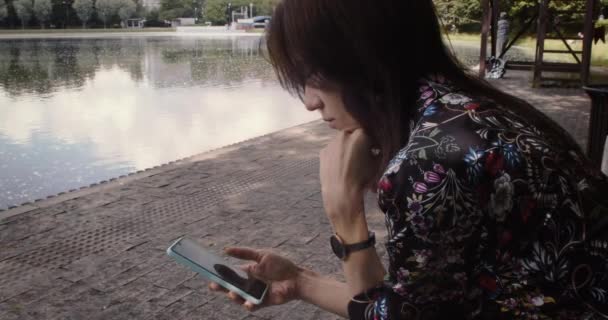 Frau sitzt im Park und nutzt ihr Smartphone, um auf soziale Netzwerke zuzugreifen — Stockvideo