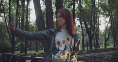 Millennial Woman sosyal ağlar için akıllı telefonunu kullanarak parkta yürürken selfie çekiyor. Her yerde bağlı kalmak