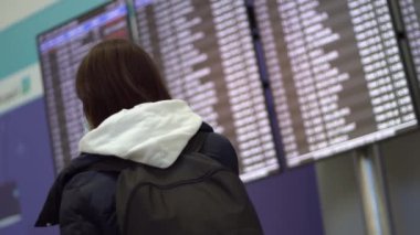 Uluslararası havaalanında uçuş bilgi panosunun yanında bir kadın var. Gezgin havaalanındaki bilgi panosuna bakar ve cep telefonuyla konuşur.