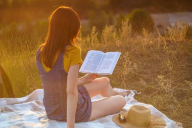 Kitabı olan genç bayan doğada dinleniyor ve gün batımında kitap okuyor, dinleniyor ve tatilin tadını çıkarıyor.