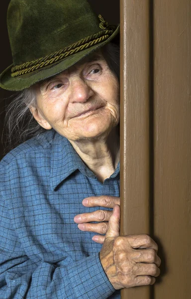 Elderly woman with hat peeking through doorway at home — Stock Photo, Image