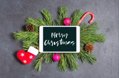 Koyu beton arka planda Noel veya yeni yıl kompozisyonu. Selamlaşmak için çam dalları ve bir tablet. Alet ekranına yazılan mutlu noeller..