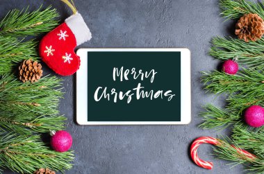 Koyu beton arka planda Noel veya yeni yıl kompozisyonu. Selamlaşmak için çam dalları ve bir tablet. Alet ekranına yazılan mutlu noeller.