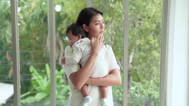 若いアジア系の母親が子供を抱いて立っている 家のリビングルームで寝るように彼を誘い出すために — ストック動画
