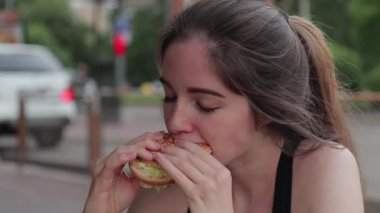 Aç bir kadın sokakta çalıştıktan sonra iştah açıcı bir şekilde hamburger çiğniyor. Kapat.