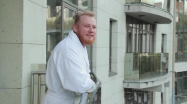Sakallı ve bornozlu genç adam tatilde bir otel balkonunda dinleniyor.