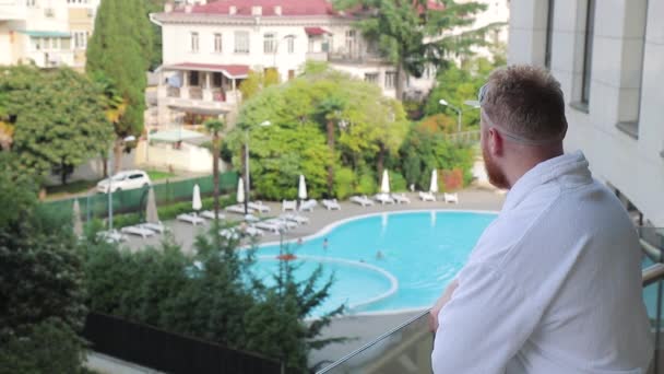 Ung mand går ud til hotellets balkon i en hvid kåbe og ser på poolen – Stock-video