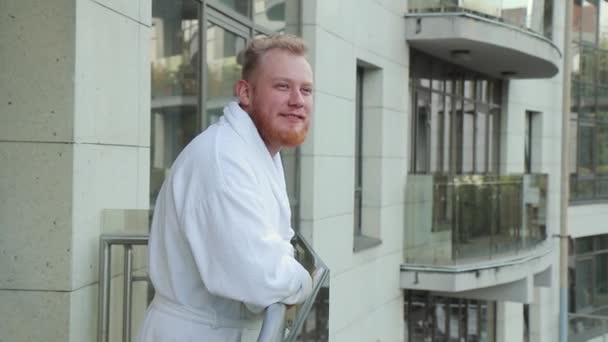 Jonge man met baard en in een badjas rust op een balkon van het hotel op vakantie — Stockvideo