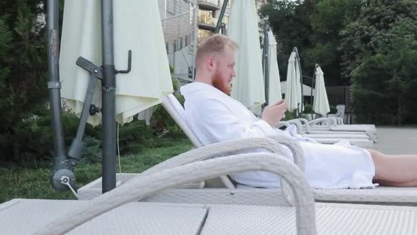 Joven con barba se sienta junto a la piscina del hotel de vacaciones con un teléfono en sus manos — Vídeos de Stock