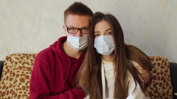 Jovem casal em máscaras médicas assistir a um filme, abraço e beijo no sofá — Vídeo de Stock