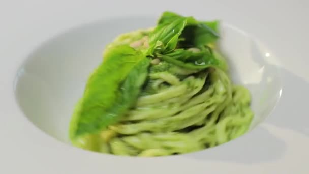 Итальянская зеленая паста с кедровыми орехами, соусом песто и свежим базиликом на белой тарелке — стоковое видео