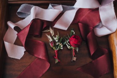 Wedding. Boutonniere. Grain. Artwork. Groom and groomsmen boutonnieres of red flowers and greens lying on a wooden tray with silk ribbons of color Marsala