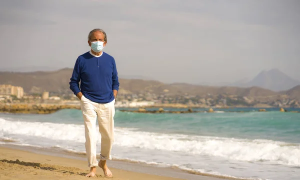 上世纪70年代的退休老人 头戴外科口罩 在海滩上散步 头戴眼罩 体贴入微地在海上散步 对健康问题进行思考 在此期间 退休老人在海滩上放松了下来 — 图库照片