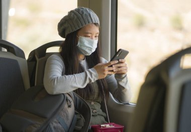 Yeni normal seyahat - çekici ve güzel Asyalı kadın yüz maskeli, tren vagonunda pencere kenarına oturmuş, trende internet cep telefonu kullanıyor.