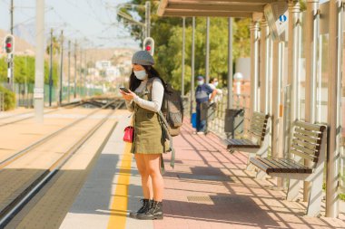 Genç, mutlu ve güzel Asyalı bir kadın, yeni normal, çekici bir Japon kız sırt çantasıyla istasyonda trenini bekliyor. Virüs turizmi gibi.