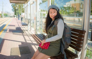 Dışarıda, mutlu ve güzel Asyalı Çinli bir turistin portresi. Sırt çantasıyla tren istasyonundaki bankta oturmuş tatil ve tatil için heyecanlı bir şekilde bekliyor.