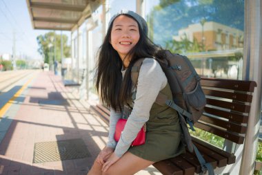 Dışarıda, mutlu ve güzel Asyalı Çinli bir turistin portresi. Sırt çantasıyla tren istasyonundaki bankta oturmuş tatil ve tatil için heyecanlı bir şekilde bekliyor.