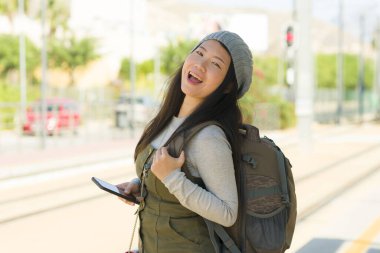 Sırt çantalı, mutlu ve güzel Asyalı Çinli kadın tren istasyonunda bekliyor. Neşeli ve heyecanlı bir şekilde gülümsüyor. Tatil seyahatinin tadını çıkarıyor.