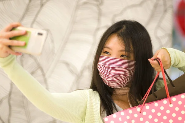 new normal at shopping mall - lifestyle portrait of young beautiful and happy Asian Korean woman in surgical mask taking selfie picture with mobile phone holding shopping bags