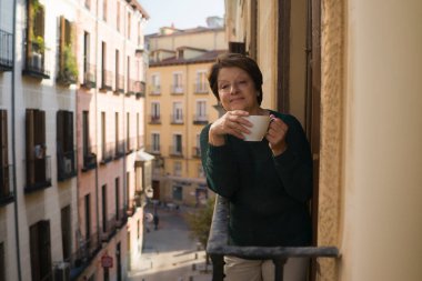 Neşeli, olgun ve sağlıklı bir kadının portresi. 60-65 yaşları arasında balkonda rahatlamış, kahve ya da çay içiyor ve sağlıklı emeklilik konseptiyle sokağa bakıyor.