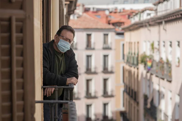 mature man in balcony home lockdown during covid19 outbreak - senior male on his 70s in face mask worried and tired looking to the street thoughtful and depressed in quarantine