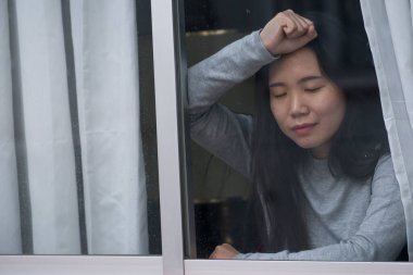 Evde stresli bir kız - üzgün ve depresif Asyalı genç bir kadının dramatik portresi. Pencerede endişeli ve çaresiz hissediyordu.