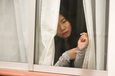 Evde stresli bir kız - üzgün ve depresif bir Asyalı kadının dramatik portresi pencerenin önünde endişeli ve umutsuz hissediyordu.