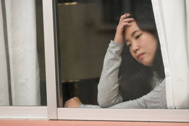 Evde stresli bir kız - üzgün ve depresif bir Asyalı kadının dramatik portresi pencerenin önünde endişeli ve umutsuz hissediyordu.