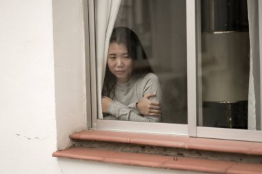 Evde stresli bir kız - üzgün ve depresif Asyalı genç bir kadının dramatik portresi. Pencerede endişeli ve çaresiz hissediyordu.