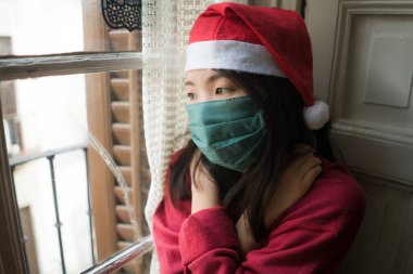 sad Christmas home alone during covid19 - young beautiful and depressed Asian Chinese woman in Santa Claus hat and face mask looking melancholic and unhappy feeling lonely clipart