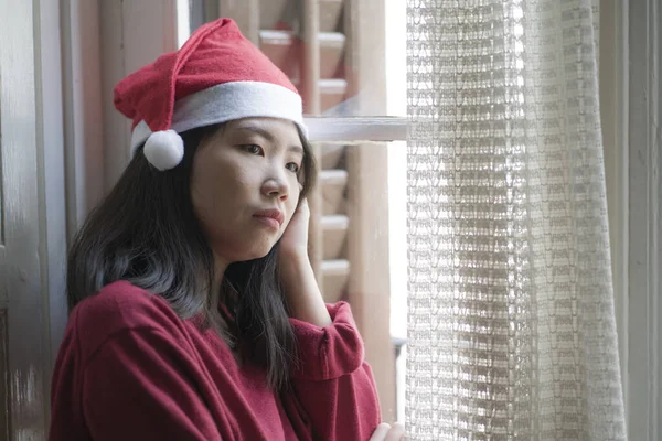 sad Christmas home alone during covid19 - young beautiful and depressed Asian Korean woman in Santa Claus hat looking melancholic through window feeling unhappy and lonely