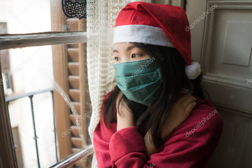sad Christmas home alone during covid19 - young beautiful and depressed Asian Chinese woman in Santa Claus hat and face mask looking melancholic and unhappy feeling lonely