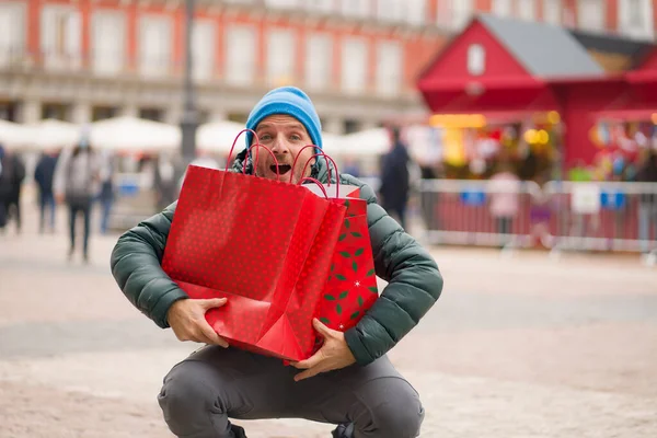 Christmas shopping in town - urban lifestyle portrait of happy and excited man in winter hat carrying shopping bags at xmas market smiling cheerful and excited buying presents