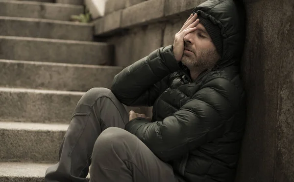 dark and edgy urban portrait of middle aged sad and depressed unemployed man sitting outdoors on dirty street corner staircase feeling upset suffering depression problem