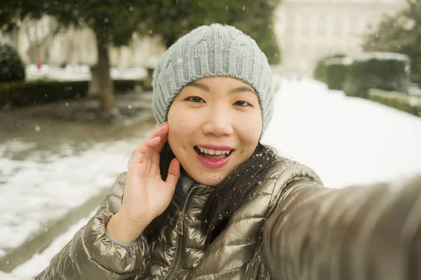 winter lifestyle portrait of young happy and beautiful Asian Korean woman taking selfie picture with mobile phone enjoying snow at city park during Christmas holidays travel