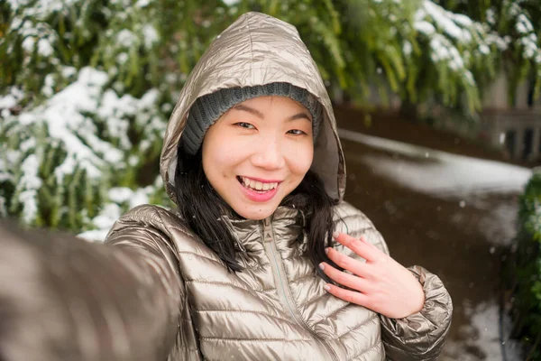 winter lifestyle portrait of young happy and beautiful Asian Korean woman taking selfie picture with mobile phone enjoying snow at city park during Christmas holidays travel