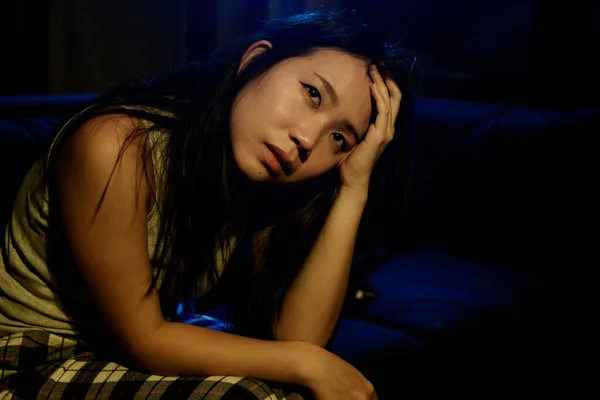 Asian woman suffering depression - dramatic artistic portrait of young beautiful sad and depressed Chinese girl in pain helpless on couch at home in the dark