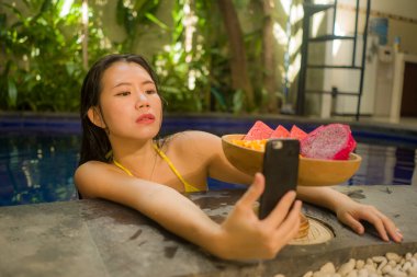 Otelde selfie. Bikinili, çekici ve mutlu Asyalı kadın. Tropikal villa havuzunda meyve tabağıyla selfie çekiyor.