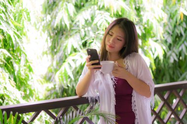 Mutlu ve güzel Asyalı bir kadının lüks otel odasının balkonunda cep telefonunda sosyal medyayı kullanarak tatilin tadını çıkarırken çekilmiş bir portresi.