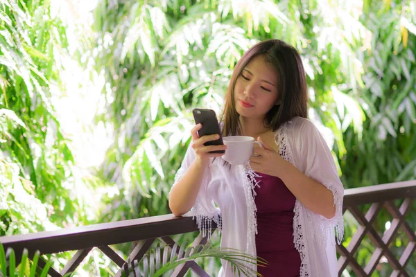 Mutlu ve güzel Asyalı bir kadının lüks otel odasının balkonunda cep telefonunda sosyal medyayı kullanarak tatilin tadını çıkarırken çekilmiş bir portresi.