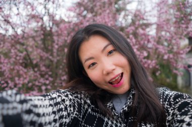 bahar sakura selfie 'si - genç mutlu ve sevimli Asyalı Koreli turist kadın cep telefonuyla kendi portresini çekiyor güzel pembe renkli ağaçların önünde neşeyle gülümsüyor