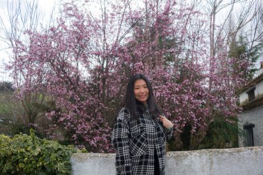 Açık havada genç mutlu ve güzel Asyalı Japon kadının portresi. Pembe çiçeklerle çiçek açan ağaçtan keyif alıyor. Neşeli gülüyor.