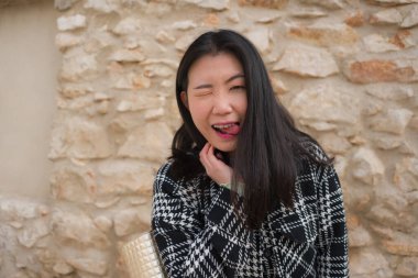 Mutlu ve güzel Asyalı bir kadının doğal portresi şık bir sonbahar ceketi içinde neşeli bir poz vererek dışarıda taş duvar arkasında pozitif titreşimler yaratıyor.