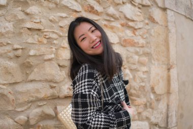 Mutlu ve güzel Asyalı bir kadının doğal portresi şık bir sonbahar ceketi içinde neşeli bir poz vererek dışarıda taş duvar arkasında pozitif titreşimler yaratıyor.