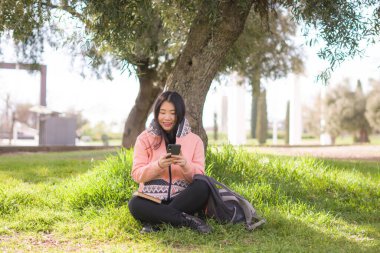 Şehir parkında cep telefonu kullanan mutlu Asyalı kadın. Yeşil çimlerin üzerinde oturan tatlı ve güzel Çinli kız. Cep telefonuyla sosyal medyanın keyfini çıkarıyor.