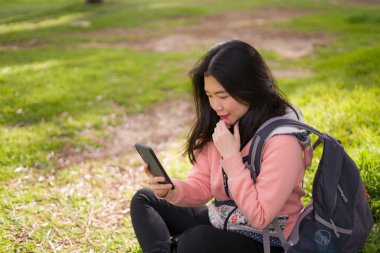 Şehir parkında cep telefonu kullanan mutlu Asyalı kadın. Yeşil çimlerin üzerinde oturan tatlı ve güzel Çinli kız. Cep telefonuyla sosyal medyanın keyfini çıkarıyor.