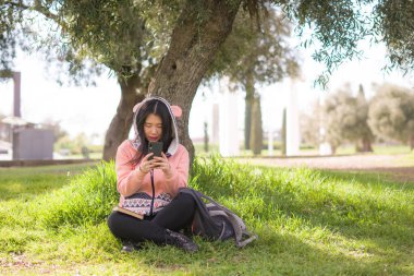 Şehir parkında cep telefonu kullanan mutlu bir Asyalı kadın. Yeşil çimlerin üzerinde oturan tatlı ve güzel bir Japon kız. Cep telefonuyla sosyal medyanın tadını çıkarıyor.